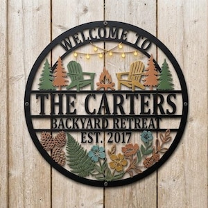 May include: A circular black metal sign featuring the text WELCOME TO THE CARTERS BACKYARD RETREAT EST. 2017. The design includes cutouts of two chairs, a campfire, string lights, pine trees, pinecones, ferns, and various colorful flowers.