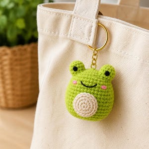 May include: A lime green crocheted frog keychain featuring a round body, two small ears, black embroidered eyes, a curved black mouth, and pink stitched cheeks. A circular beige patch is centered on the belly, attached to a gold metal ring.