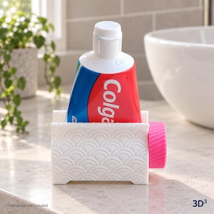 May include: A white plastic toothpaste tube squeezer featuring a repeating embossed wave pattern on the front. The device has a bright pink circular knob on the side and holds a tube of Colgate toothpaste with a white cap in an upright position.