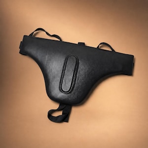 May include: A black protective groin guard featuring a contoured, padded central section with a smooth surface. The gear includes adjustable black elastic straps at the top and bottom to secure the piece in place during physical activity.