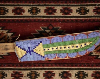 May include: A knife sheath featuring intricate beadwork in light blue, green, and yellow. The design includes a central green stripe with dark blue accents, bordered by light blue sections with yellow geometric patterns on a wooden base.