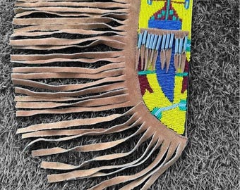 May include: A tan suede leather cuff featuring a rectangular panel of intricate beadwork in yellow, blue, red, and turquoise. The cuff is adorned with long, thin suede fringe strips and a row of silver-toned metal cones hanging from the beaded section.