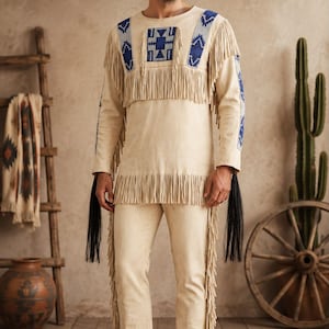 May include: A cream-colored long-sleeve shirt and matching pants set featuring long fringe detailing along the chest, sleeves, and legs. The shirt includes blue and white geometric patterns on the shoulders and chest, with long black tassels.