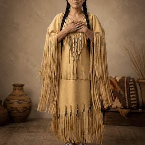 May include: A tan suede dress featuring long fringe detailing along the sleeves and hemline. The garment includes a chest panel adorned with vertical rows of silver-toned metal beads and matching fringe accents. The outfit is paired with beaded moccasins.