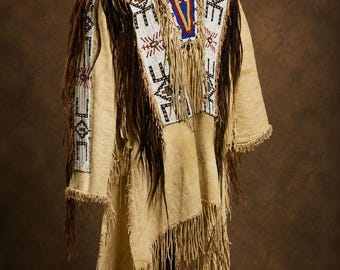 May include: A tan buckskin tunic featuring extensive fringe detailing along the sleeves and hem. The garment is adorned with intricate white, red, blue, and green beadwork patterns across the chest and shoulders, complemented by long dark hair fringe.
