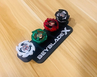 May include: A black display stand featuring the white text BEYBLADE X, holding three spinning top toys. The tops are arranged in a row, colored white, green, and red, with a fourth black and white top positioned at the end of the display base.
