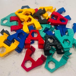 May include: A collection of small, multi-colored plastic clips in red, yellow, blue, black, and teal. Each clip features a distinct hexagonal honeycomb texture on its surface and a U-shaped opening designed for securing or organizing items.