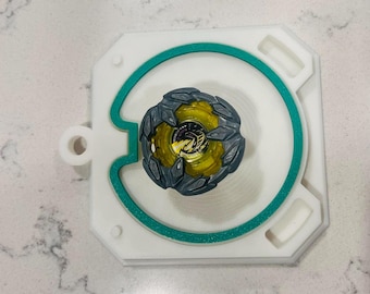 May include: A circular spinning top toy with a translucent yellow center and a dark gray, jagged outer ring, resting on a white square platform featuring a teal circular inlay and a small circular cutout on the left side of the frame.