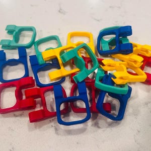 May include: A collection of small, U-shaped plastic clips in vibrant primary and secondary colors including red, blue, yellow, and teal. Each clip features a square-like frame with a hinged mechanism, arranged in a loose, overlapping pile.