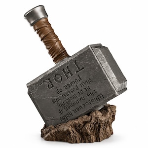 Op de afbeelding: Een replica van Thor's hamer, Mjolnir, met een verweerde grijze metalen kop met een gebarsten steentextuur en de inscriptie: Whosoever holds this hammer, if he be worthy, shall possess the power of THOR. De hamer heeft een bruin, met leer omwikkeld handvat en rust op een bruine rotsachtige basis.