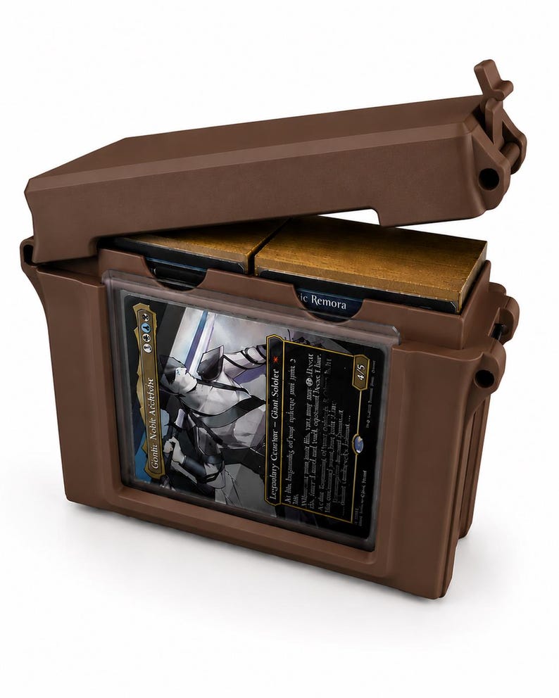 May include: A brown plastic deck box with a hinged lid, featuring a clear front display window holding a Gonti, Cabel Archfiend trading card. The interior contains two stacks of cards with gold-colored edges visible at the top of the container.