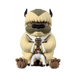 Op de afbeelding: Een vinylfiguur van een groot, harig, crèmekleurig wezen met bruine markeringen, zwarte hoorns en een donkerbruine neus, dat een kleiner, wit en bruin gevleugeld wezen met grote groene ogen zittend tegen de borst houdt.