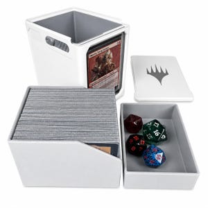 Könnte beinhalten: Eine weiße, modulare Kartenaufbewahrungsbox mit grauem Innenraum, die einen Stapel Sammelkarten enthält und ein Sichtfenster mit einer Daretti, Scrap Savant-Karte aufweist. Enthält ein separates graues Fach mit vier Würfeln und einen weißen Deckel.