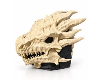 May include: A cream-colored dragon skull sculpture featuring prominent horns, sharp teeth, and detailed bone-like textures. The skull is mounted on a solid black base with a segmented, blocky design that contrasts with the organic shape.