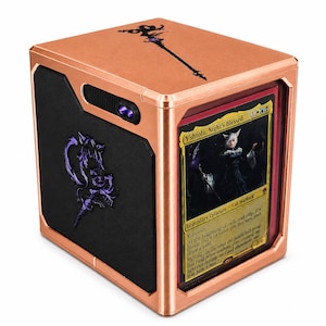 May include: A copper-colored metal deck box featuring a black side panel with a purple emblem and a top surface with a black staff graphic. The front displays a Y'shtola, Night's Blessed trading card behind a clear protective window.