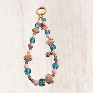 May include: A beaded keychain featuring a gold-toned metal ring at the top. The loop is composed of alternating light pink round beads, textured turquoise crackle glass beads, and small metallic gold-toned seashell charms arranged in a sequence.