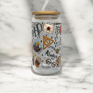 May include: A clear glass tumbler with a bamboo lid and a glass straw, featuring a wrap-around design of various fantasy-themed illustrations including a castle, a golden snitch, round glasses, a letter, and the text Always and Muggles.