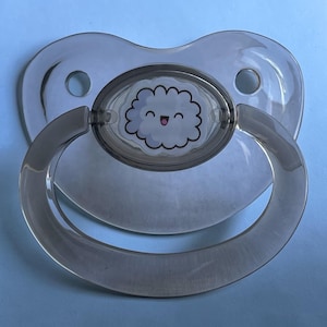 May include: A clear, transparent pacifier featuring a central oval emblem decorated with a cartoon illustration of a smiling, fluffy white cloud with a pink mouth. The pacifier has a curved handle and a shield with two circular ventilation holes.