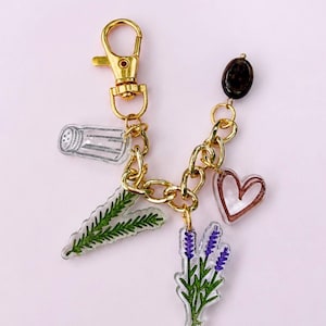 May include: A gold-toned metal keychain featuring a lobster clasp and a thick chain. Attached are five charms: a clear salt shaker, a green rosemary sprig, a purple lavender sprig, a pink-outlined heart, and a single dark brown oval bead.