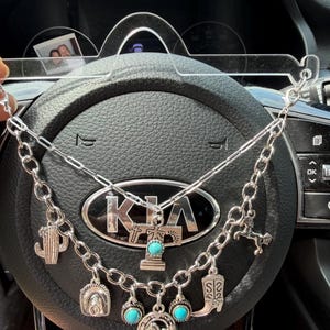May include: A silver-toned charm necklace featuring a layered chain design. The charms include a cactus, a tombstone with a turquoise stone, a cowboy hat, a horseshoe with a horse, and a cowboy boot, all finished in a metallic silver tone.
