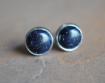 May include: A pair of round stud earrings featuring dark blue, glittery, star-like flecks set within a smooth, polished silver-toned metal frame. The circular stones have a deep navy hue with reflective, sparkling inclusions that mimic a night sky.