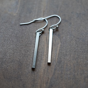 May include: A pair of minimalist silver bar earrings. Each earring features a straight, rectangular bar suspended from a simple hook. The earrings are displayed on a dark brown wooden surface.