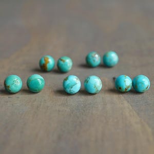 May include: Several pairs of turquoise stud earrings are arranged on a brown wooden surface. The earrings are round, with a mottled pattern of blue-green and brown. The earrings are a natural stone.