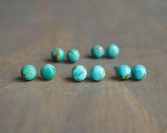 May include: Several pairs of turquoise stud earrings are arranged on a brown wooden surface. The earrings are round, with a mottled pattern of blue-green and brown. The earrings are a natural stone.