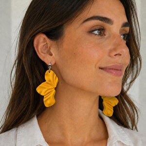 Puede incluir: Un par de pendientes colgantes de color amarillo mostaza con un diseño de abanico plisado. Cada pendiente cuelga de un pequeño perno de metal plateado. La superficie texturizada y ondulada del material amarillo crea un aspecto estratificado.