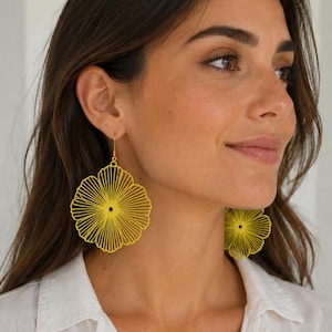 Puede incluir: Un par de pendientes colgantes de color amarillo brillante con un diseño floral circular grande y calado, con líneas finas y radiantes que se extienden desde un punto central. Los pendientes cuelgan de ganchos dorados y tienen un acabado mate.