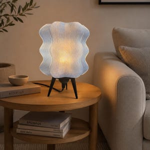Puede incluir: Una lámpara de mesa con una pantalla ondulada, acanalada y de color azul claro translúcido, con una base de trípode negra. La lámpara emite un brillo cálido y suave desde el interior, iluminando la superficie texturizada de la pantalla.