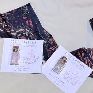 Puede incluir: Dos pequeños frascos de vidrio con tapones de corcho, cada uno sobre una tarjeta blanca con la etiqueta COZY CRYSTALS. Un frasco contiene piedras de cuarzo rosa y el otro piedras de cuarzo transparente. Cada tarjeta tiene un dibujo de cristales.