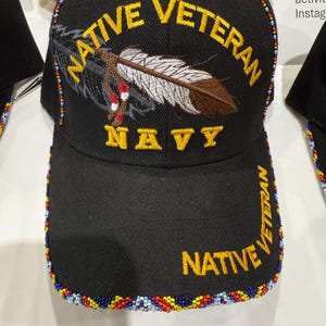 May include: A black baseball cap featuring the embroidered text NATIVE VETERAN and NAVY in yellow. An embroidered feather graphic sits in the center. The brim and crown edges are decorated with a multi-colored beaded trim in red, blue, and white.