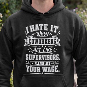 May include: A black hooded sweatshirt featuring white text that reads I HATE IT WHEN COWORKERS ACT LIKE SUPERVISORS. PLEASE ACT YOUR WAGE. The design includes decorative banners and stars surrounding the text, centered on the front of the garment.