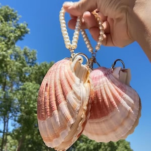 Könnte beinhalten: Zwei Jakobsmuscheln mit rosa und weißen Rillen, die jeweils an einem goldfarbenen Metallring und einer Schlaufe aus cremefarbenen Kunstperlen befestigt sind. Die Muscheln werden zusammengehalten und zeigen ihre natürlichen Texturen.