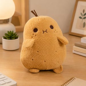May include: A plush toy shaped like a potato with a soft, golden-brown textured surface. It features two dark brown embroidered eyes, a small stitched mouth, and a single brown stem on top. The potato has two small arms and two feet.
