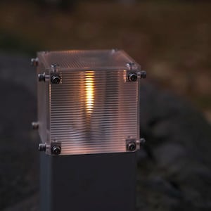 Może przedstawiać: Kwadratowa lampa zewnętrzna z przezroczystą, poziomo żebrowaną obudową z akrylu, mocowaną srebrnymi metalowymi śrubami w każdym rogu. Wnętrze emituje ciepłe światło, a podstawę stanowi ciemnoszary, prostokątny słupek.