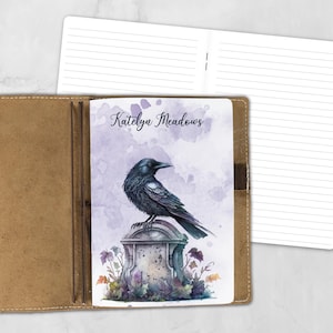 May include: A tan leather notebook cover holding a softcover journal with a watercolor illustration of a black raven perched on a tombstone. The text Katelyn Meadows is printed above the raven. An open, lined notebook is visible behind.