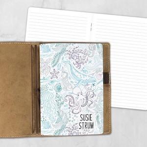May include: A tan leather notebook cover holding a white insert with a blue and purple line art pattern of sea creatures including an octopus, whale, crab, and starfish. The text SUSIE STRUM is printed in black on the lower right corner.