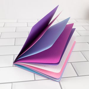 May include: A notebook with multiple pages fanned open, featuring a gradient of colors including deep purple, light blue, magenta, white, and soft pink. Each page is made of paper with a subtle dot grid pattern visible across the surface.