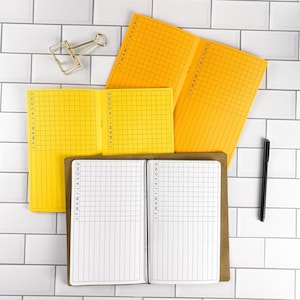 May include: Three open notebooks with grid paper and monthly tracking columns labeled J F M A M J J A S O N D. Two notebooks have bright yellow covers, and one has a brown cover. A gold metal binder clip and a black pen are positioned nearby.
