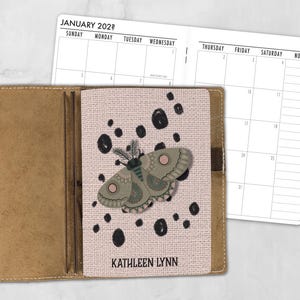 May include: A tan leather notebook cover holding a planner insert with a light pink cover featuring a moth illustration, black polka dots, and the text KATHLEEN LYNN. The open planner shows a January calendar grid with days of the week printed.