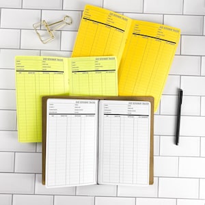 May include: Three debt repayment tracker booklets with yellow and white pages featuring columns for date, amount paid, new balance, and additional information. A gold metal binder clip and a black pen are positioned near the open booklets.