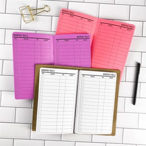 May include: Three open monthly bill trackers with grid layouts. One is white with black text, one is purple with black text, and one is bright pink with black text. Each page features columns labeled Paid, Company, Due, and Amount. A gold binder clip and a black pen are present.