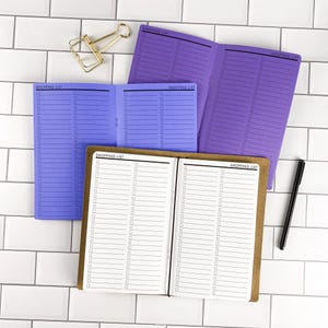 May include: Two open purple shopping list notebooks with lined pages and checkboxes, one open brown notebook with white pages featuring black lines and checkboxes, a gold metal binder clip, and a black pen on a white tiled surface.