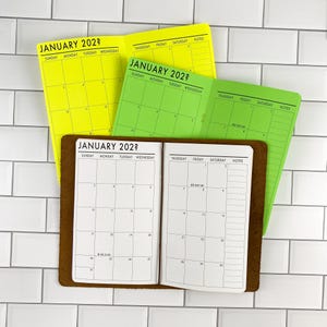 May include: Three open planners with a monthly calendar layout for January 202?. The top planner is neon yellow, the middle is lime green, and the bottom is white with a brown leather cover. Each page features a grid with days of the week.