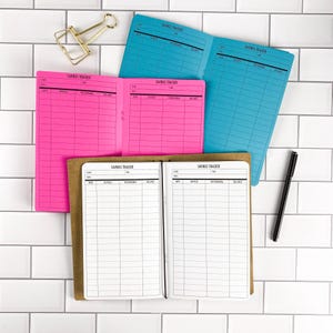 May include: Three open savings tracker booklets in bright pink, vibrant blue, and tan with white paper inserts. Each booklet features printed columns for date, deposit, withdrawal, and balance. A gold metal binder clip and a black pen are included.