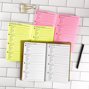 May include: Three open password keeper booklets in neon yellow, bright pink, and brown with white pages. Each booklet features printed lines for website, app, username, email, password, security question, and notes. A gold binder clip and black pen.