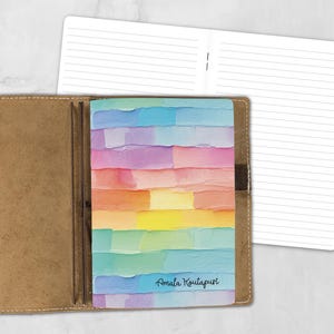 May include: A tan leather notebook cover holding a softcover journal with a colorful, textured, horizontal brushstroke pattern in pastel shades of blue, pink, yellow, and green. The text Amala Koutapuri is printed in black on the bottom right.