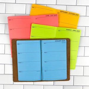 May include: A set of four paper planners in bright neon colors including yellow, pink, lime green, and blue. Each planner features printed black text for months and days of the week. One blue planner is open and inserted into a brown leather cover.